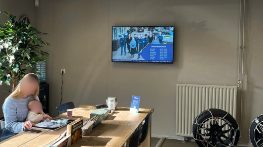 Klant aan tafel in wachtruimte met narrowcasting scherm aan de muur.